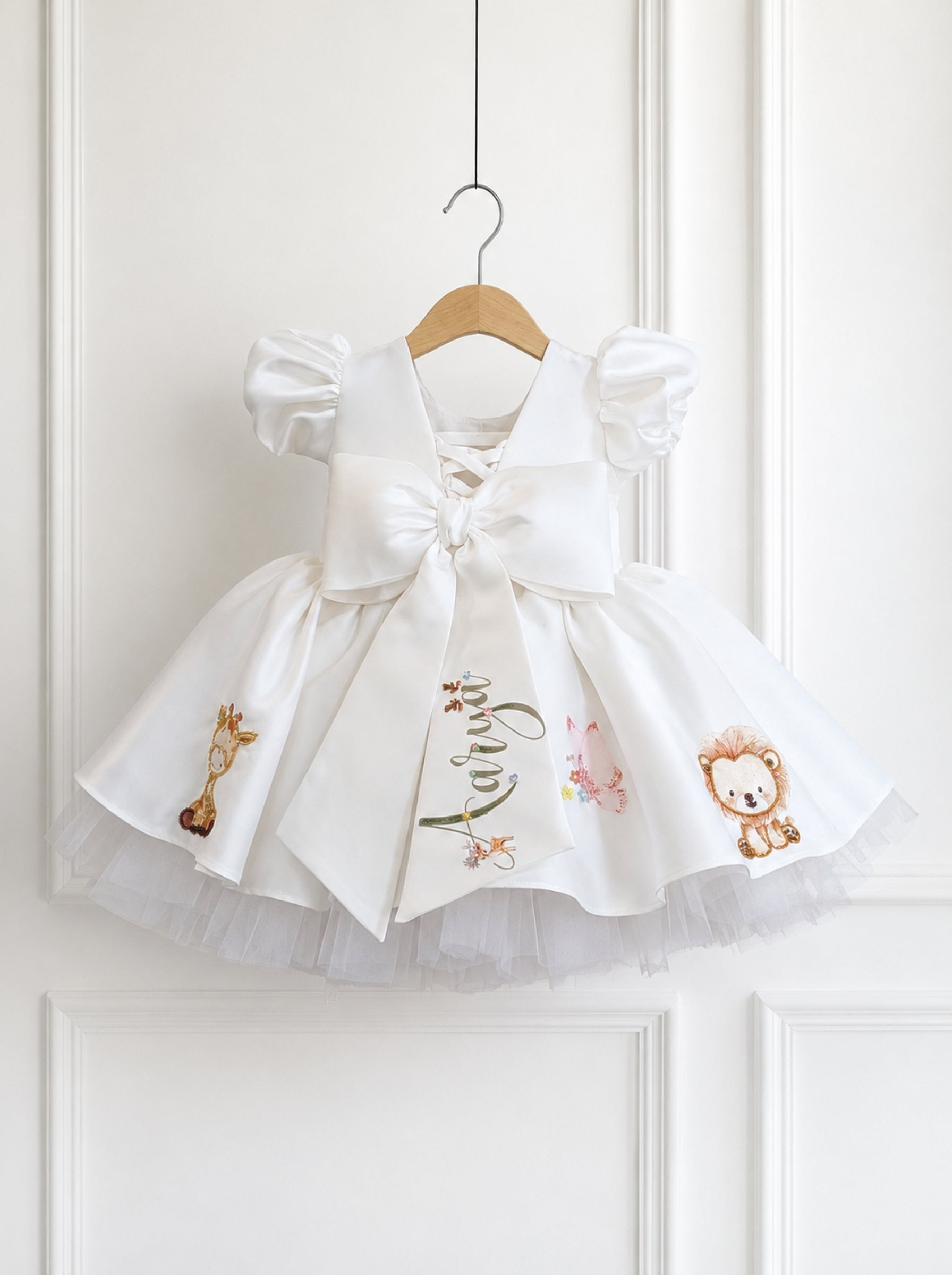 A white baby girl's dress with a large bow, puffed sleeves, and embroidered safari-themed characters including a giraffe, lion, and elephant, hanging on a wooden hanger against a white paneled door.