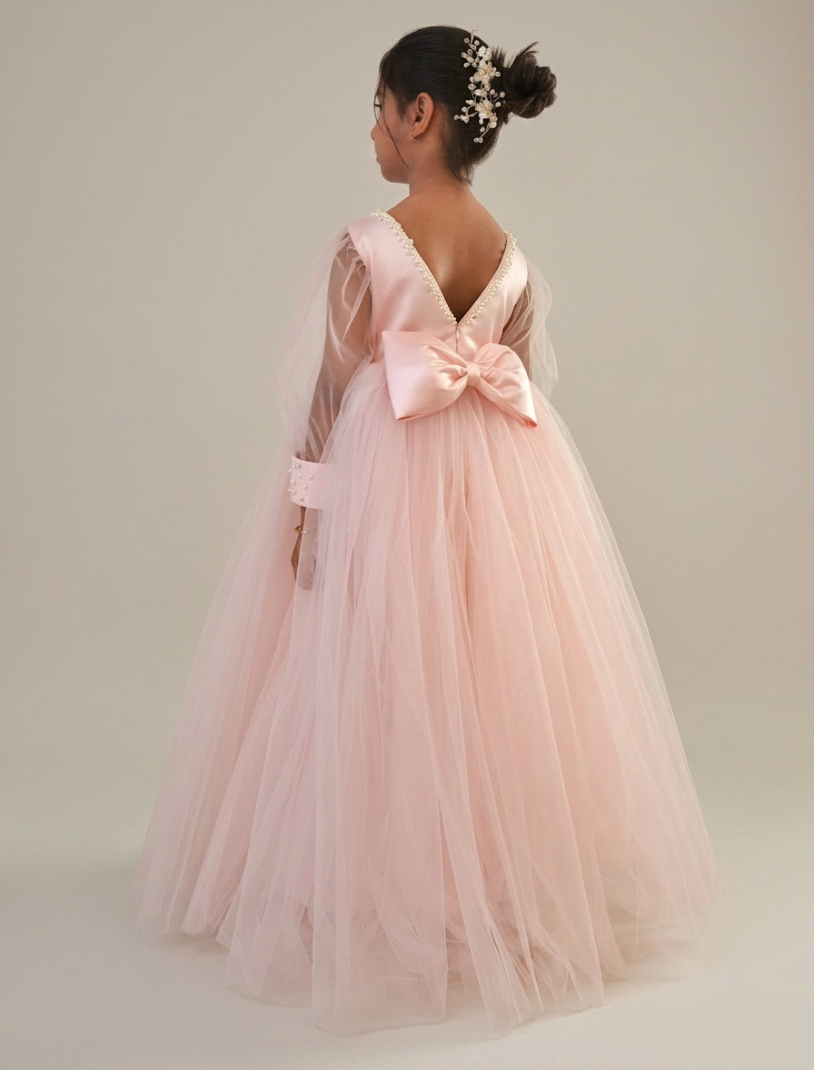 A young girl viewed from behind wearing a light pink tulle ball gown with sheer long sleeves, a deep V back, and a large matching satin bow at the back. She has her hair in a bun with a decorative hair clip, set against a plain light gray studio background.