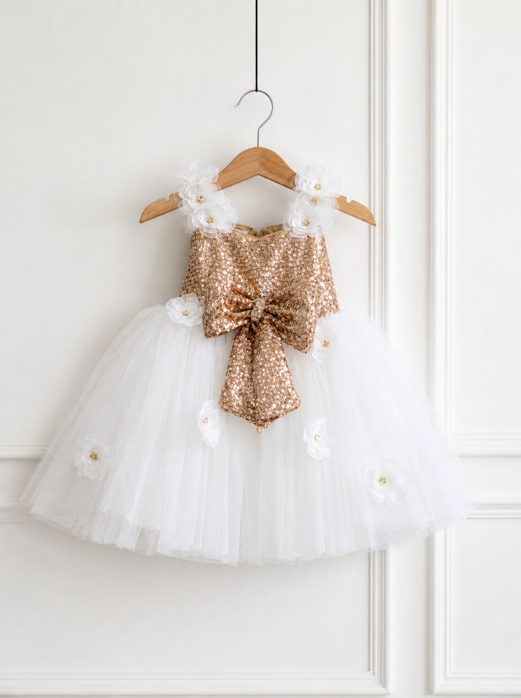 A toddler girl's party dress with a white tulle skirt and a gold sequined bodice, decorated with white fabric flowers, hanging on a wooden hanger against a white paneled wall.