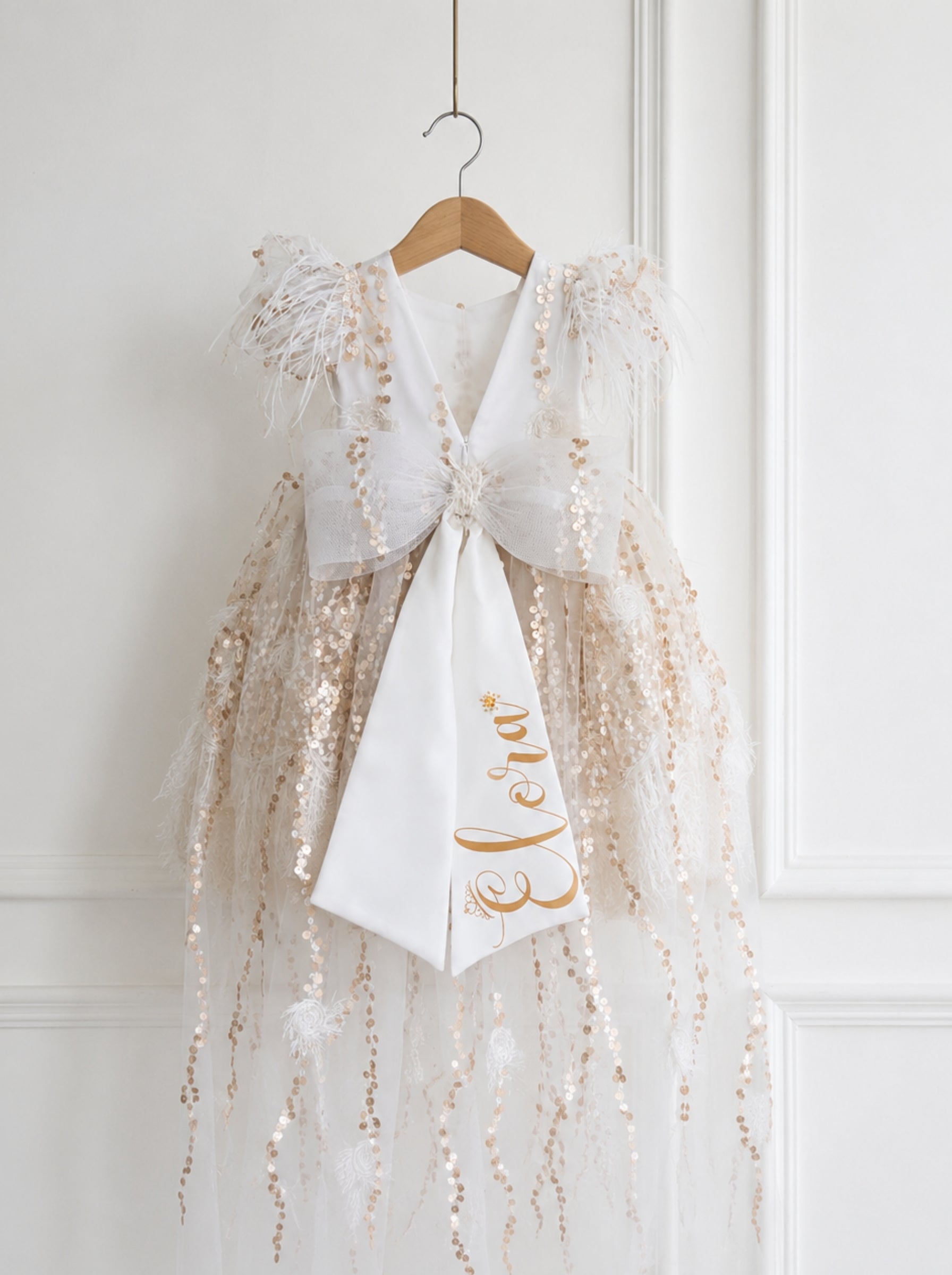 A white and gold sequined dress with a large bow and a white sash printed with the name 'Léa' in gold script, hanging on a wooden hanger against a white paneled wall.