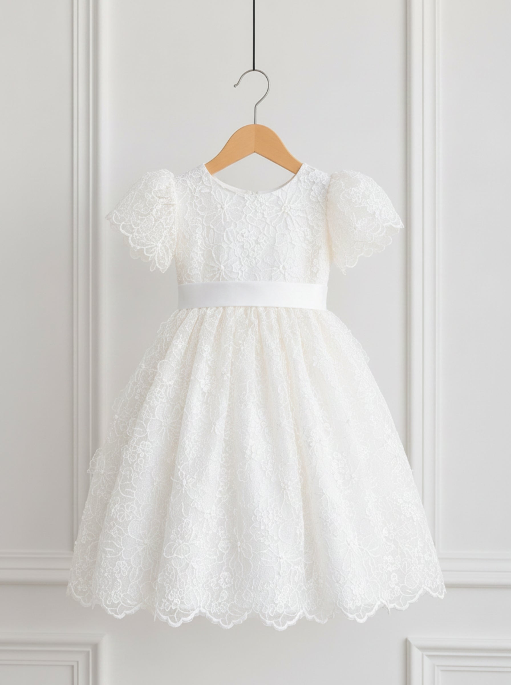 A white lace girl's party dress with short puff sleeves and a matching waistband, hanging on a wooden hanger against a white paneled wall.