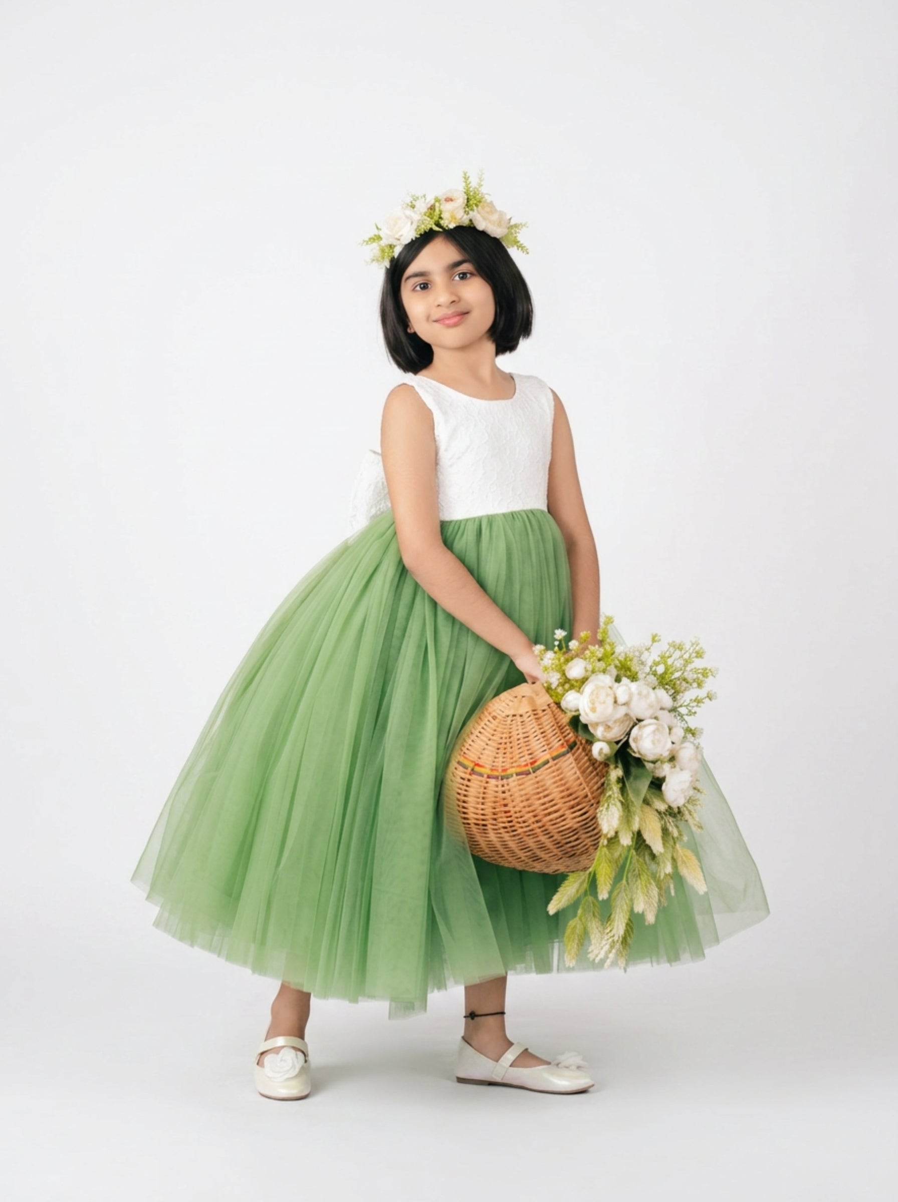 Child wearing green tulle flower girl dress with white lace bodice, floral crown, and basket of roses — luxury party wear India.