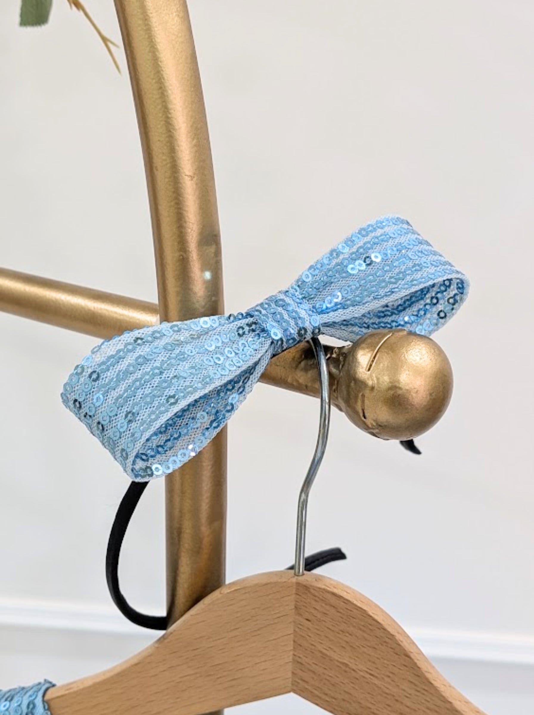 A light blue sequined fabric bow tied around a gold hanger stand, with a wooden hanger hanging below and a neutral indoor background.