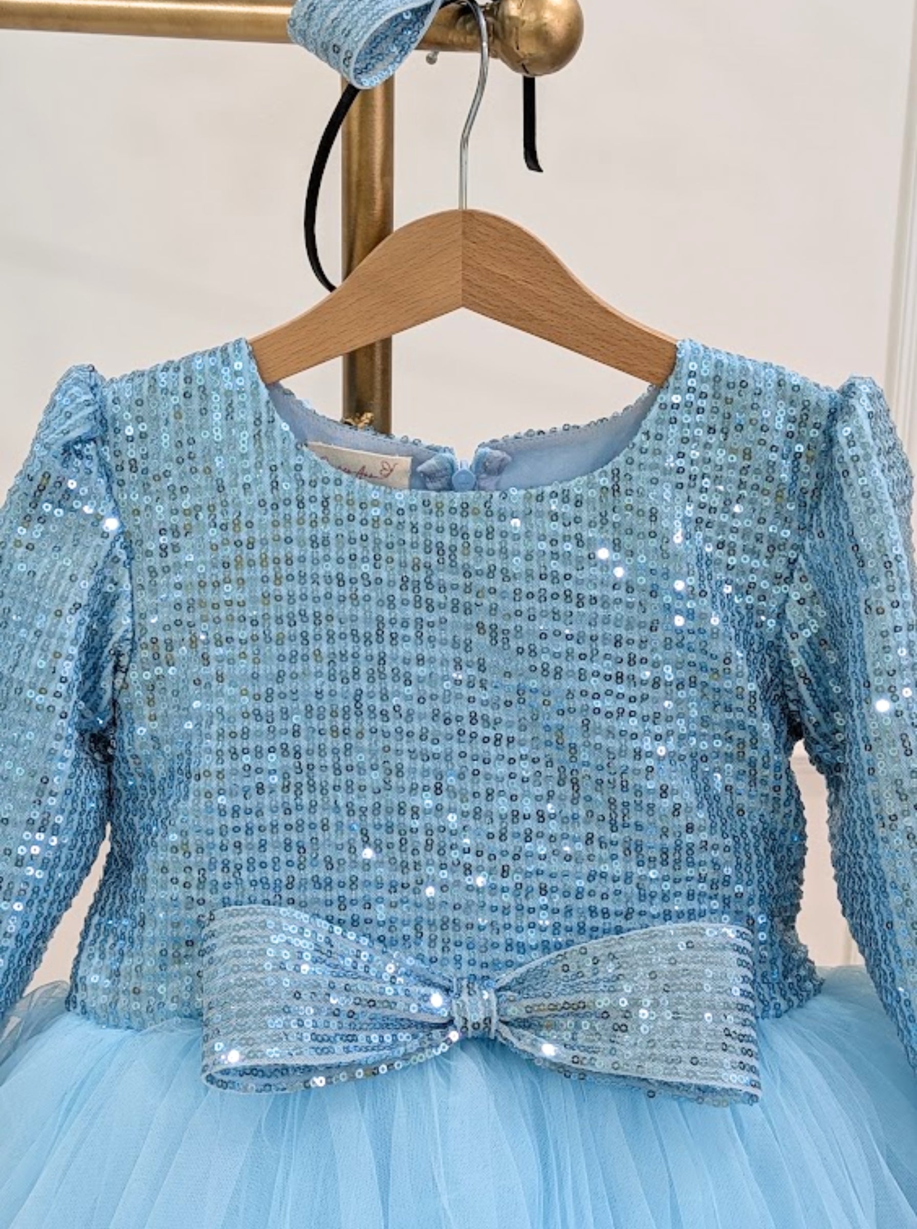 A light blue sequined girls' dress hanging on a wooden hanger, featuring a sparkling sequin bodice, long sleeves, and a matching sequined bow at the waist, with a matching headband also visible.