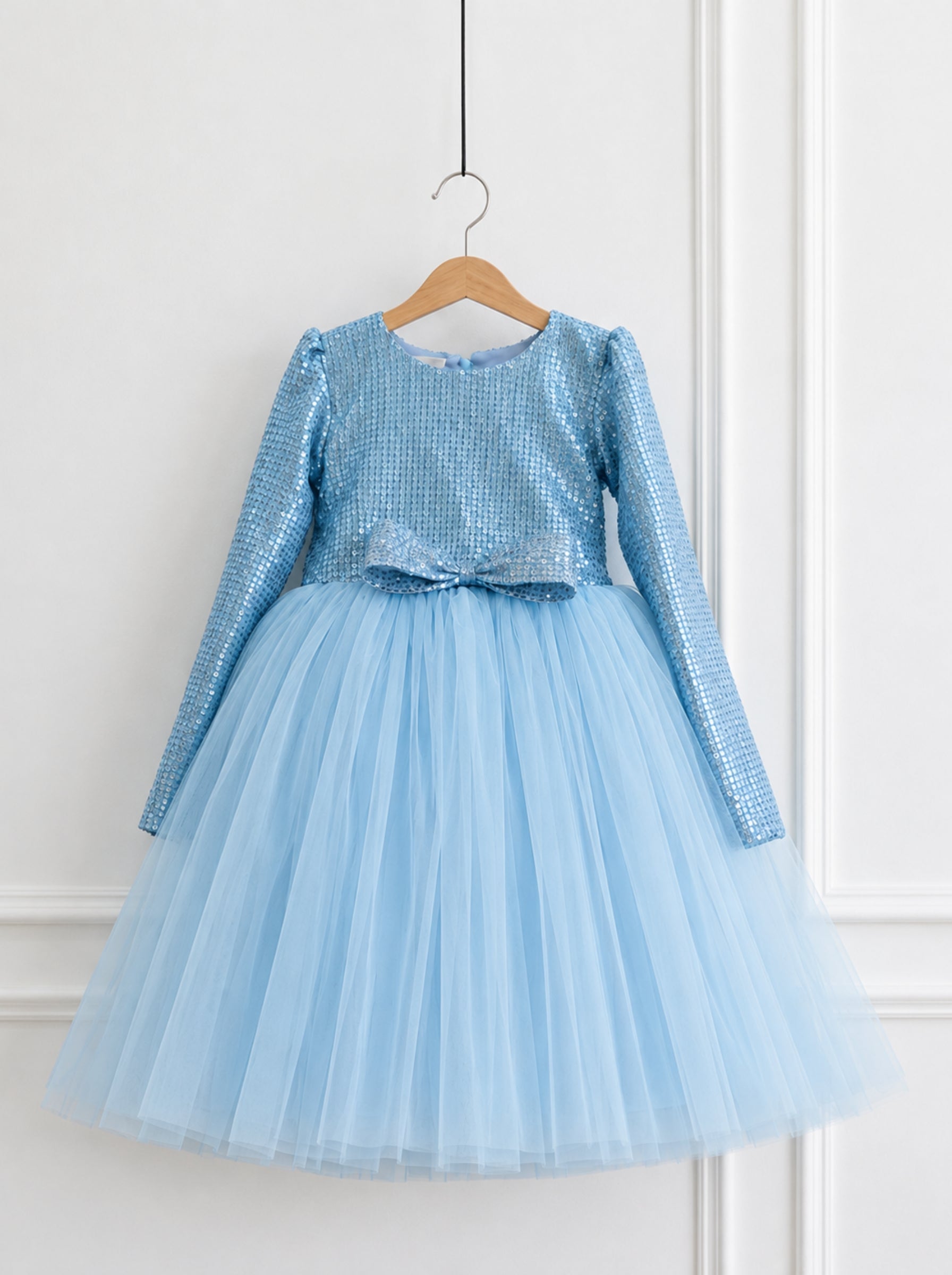 A light blue long-sleeve girl's party dress with a sequined bodice and a voluminous tulle skirt, hanging on a wooden hanger against a white paneled wall.