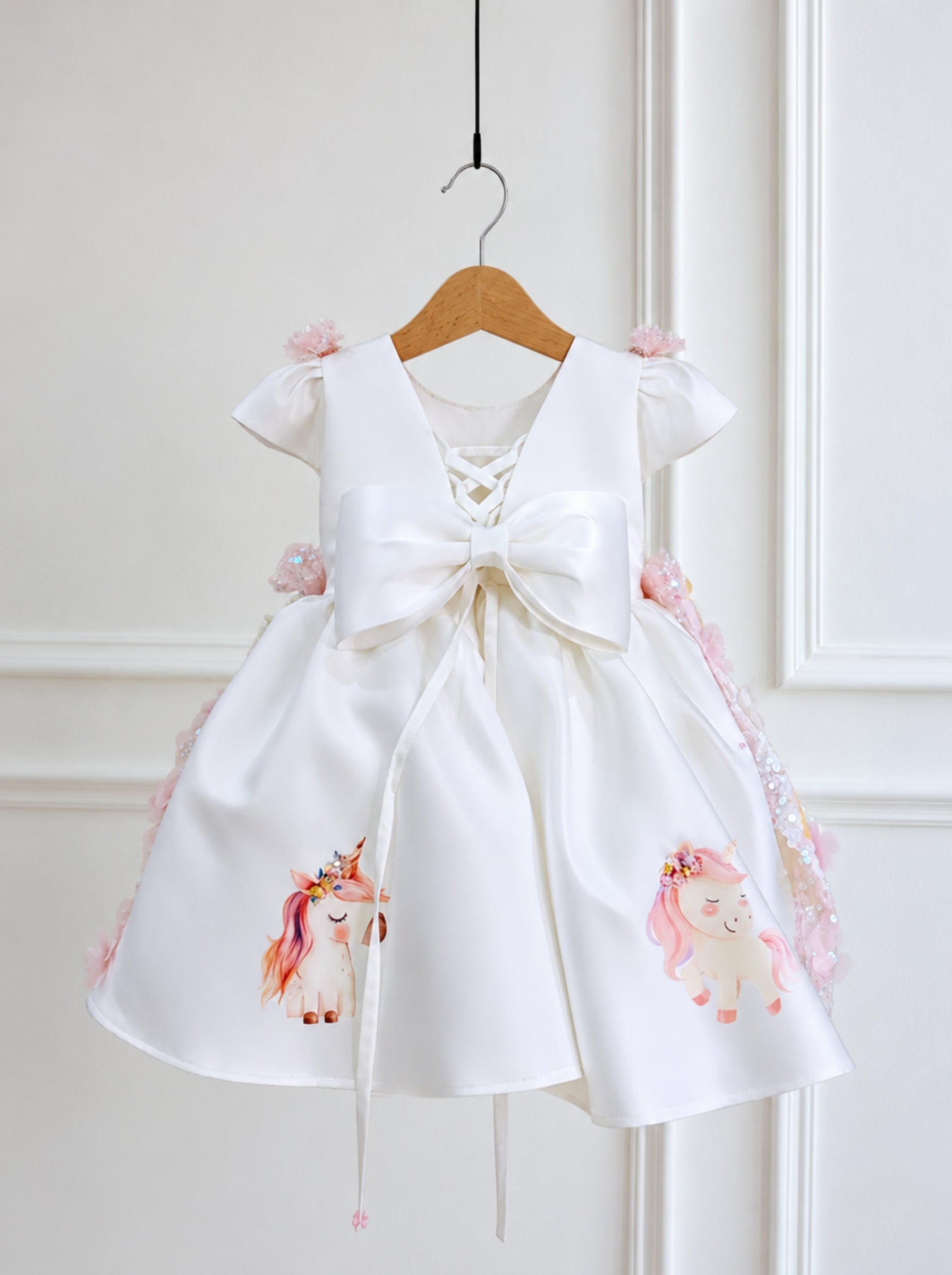 A white baby girl's dress with a large satin bow at the front, decorated with pink floral details and two watercolor unicorn prints on the skirt, hanging on a wooden hanger against a white paneled wall.