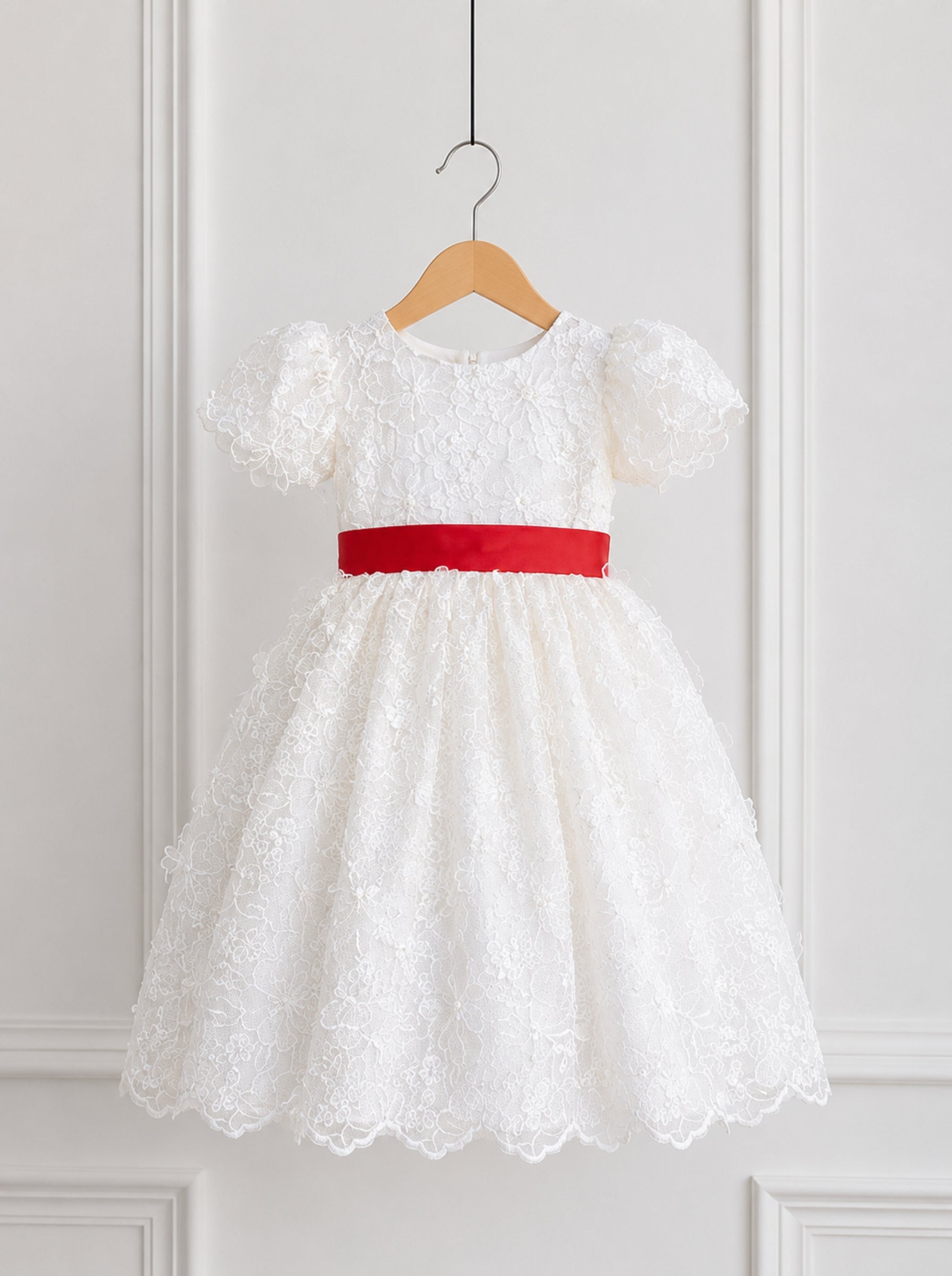 A white lace toddler girl's dress with short puff sleeves and a bright red satin waist sash, hanging on a wooden hanger against a white paneled wall.