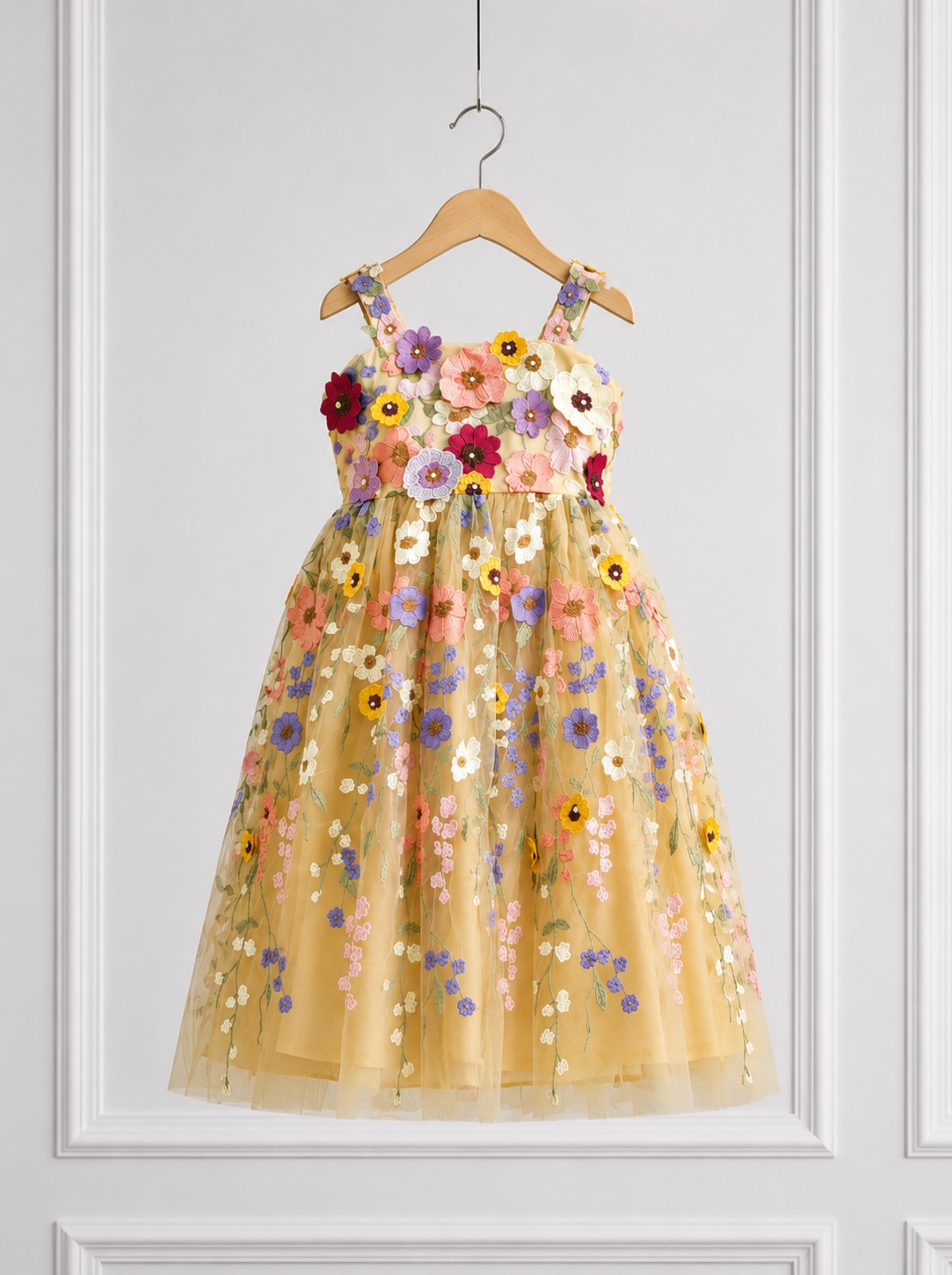 A sleeveless yellow tulle dress with colorful floral embroidery and applique, hanging on a wooden hanger against a white paneled wall.
