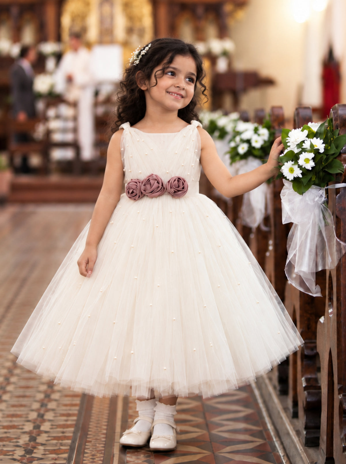 A young girl stands in a church aisle wearing a sleeveless cream tulle flower girl dress with a waistband of three dusty mauve fabric roses. 