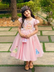 A young girl wearing a pink princess-themed dress with a carriage and character illustrations, holding a matching small bag.