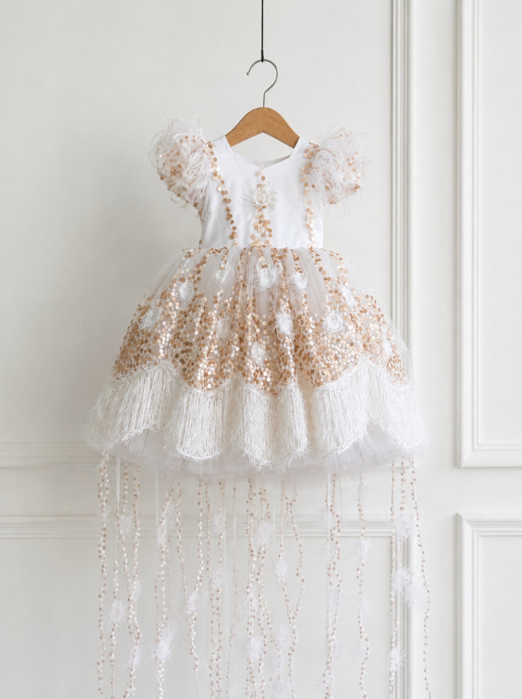 A white and gold beaded baby girl's party dress with short puff sleeves and a long tulle train, hanging on a wooden hanger against a white paneled wall.