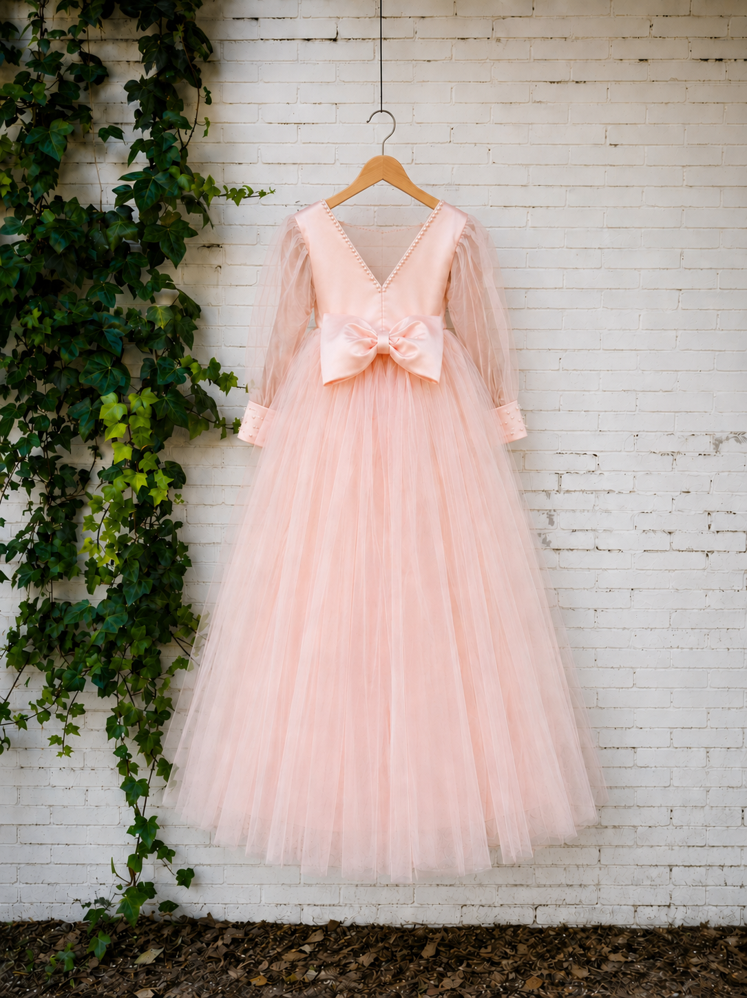 A blush pink tulle dress with a large bow at the waist and sheer long sleeves, hanging on a wooden hanger against a white brick wall with green ivy on the left.