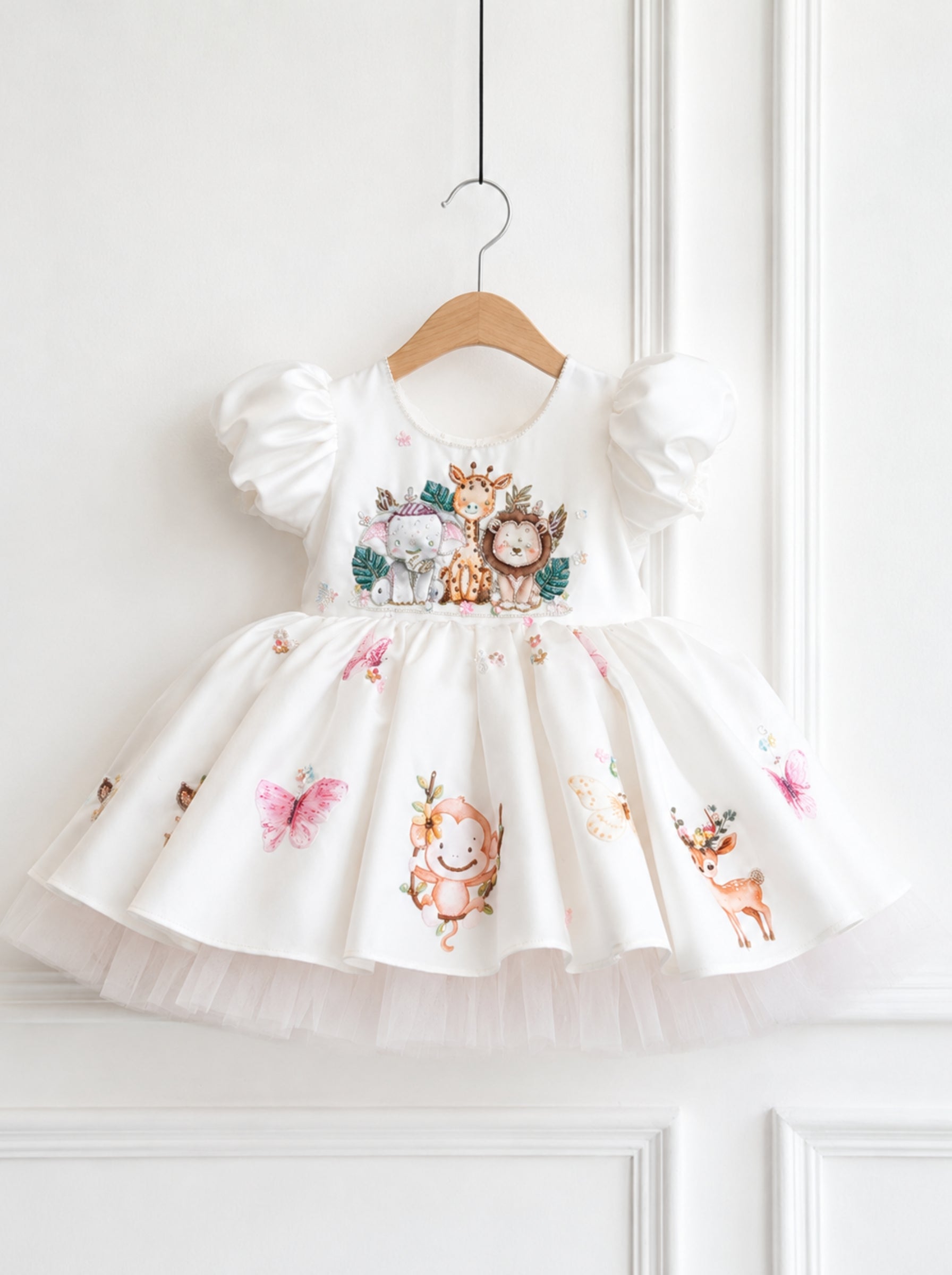 A white toddler girl's dress with puff sleeves and a tulle skirt, decorated with cute watercolor-style woodland animal prints including a bear, owl, fox, butterfly, deer, and a clock character. The dress is hanging on a wooden hanger against a white paneled wall.