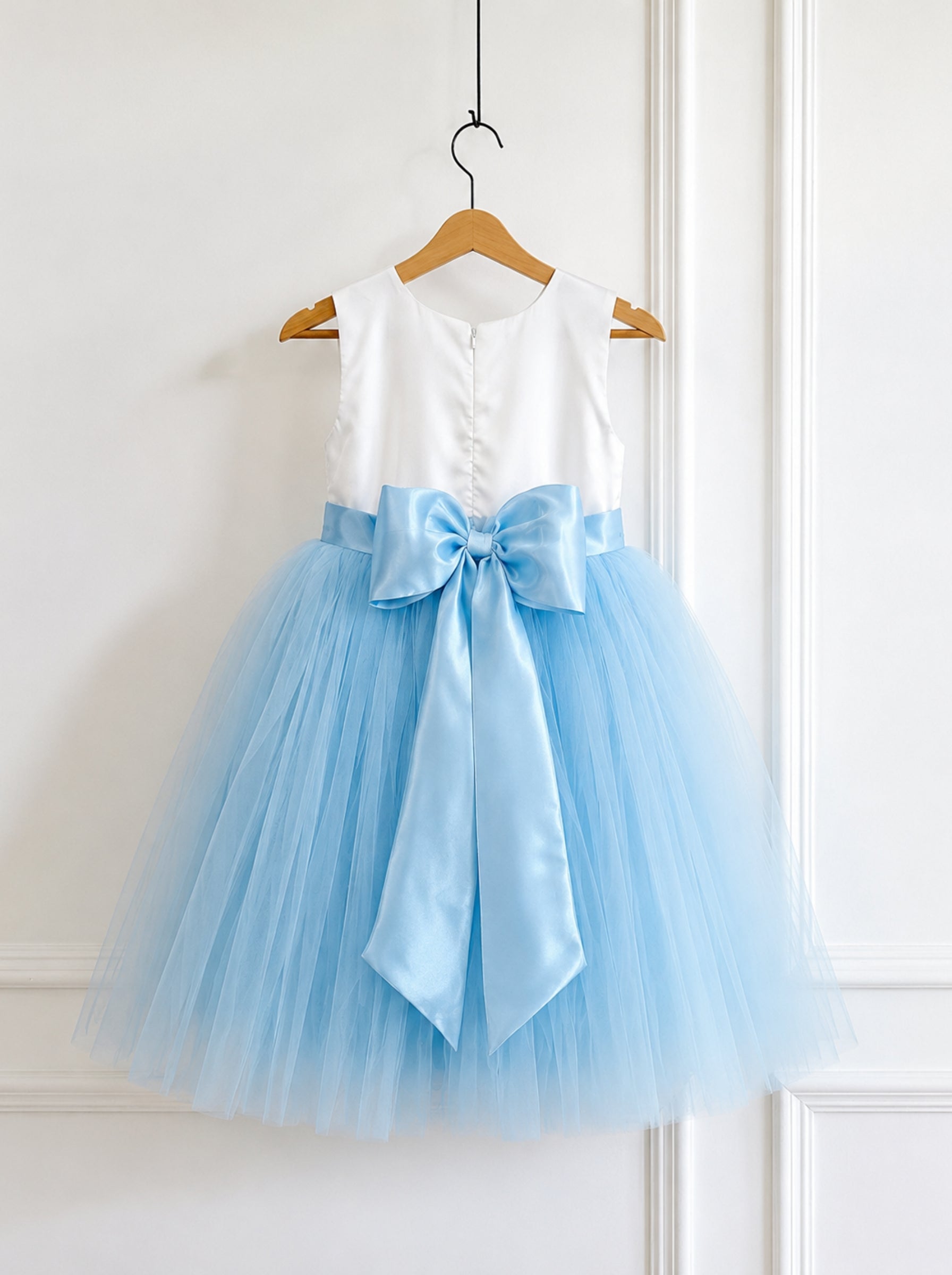 A sleeveless children's party dress hanging on a wooden hanger against a white paneled wall. The dress has a white satin bodice, a light blue satin waistband with a large bow, and a voluminous light blue tulle skirt.