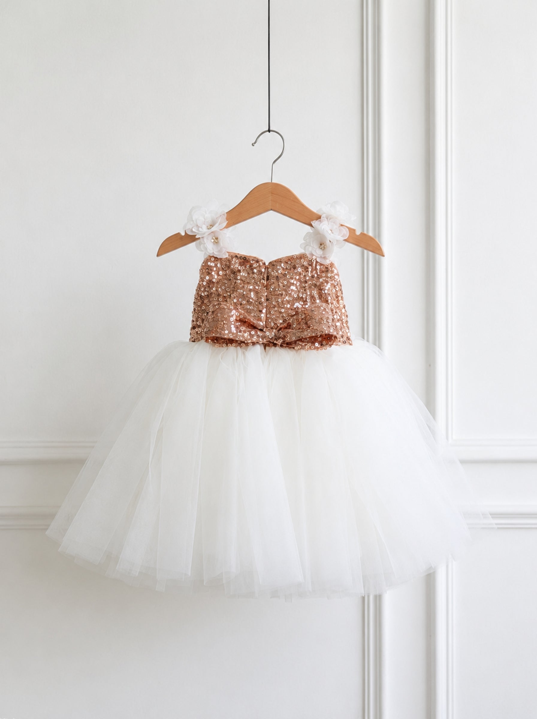 A toddler girl's strapless sequined dress hanging on a wooden hanger against a white paneled wall. The dress features a rose gold sequined bodice with white fabric flowers on the shoulders and a voluminous white tulle skirt.
