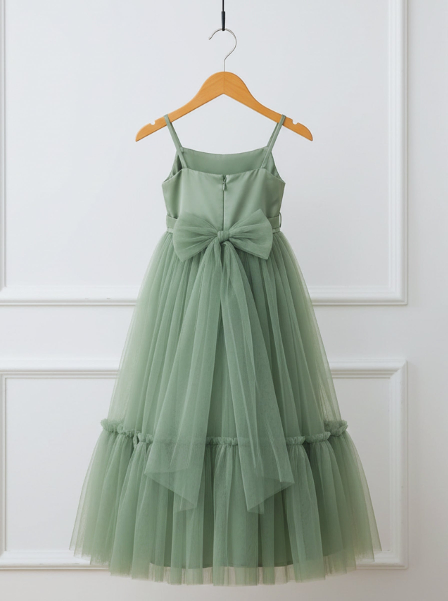 A sage green sleeveless tulle dress with spaghetti straps, a large bow at the waist, and tiered ruffle hem, hanging on a wooden hanger against a white paneled wall.