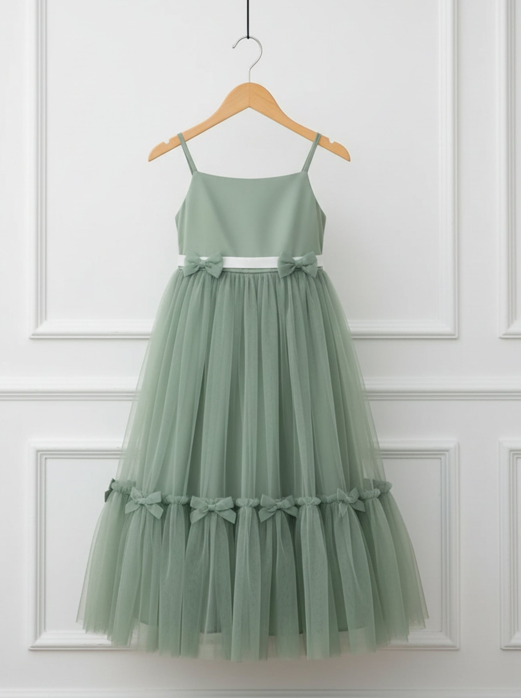 A sage green sleeveless tulle dress with thin shoulder straps, a fitted bodice, and layered ruffle tiers decorated with small bows, hanging on a wooden hanger against a white paneled wall.