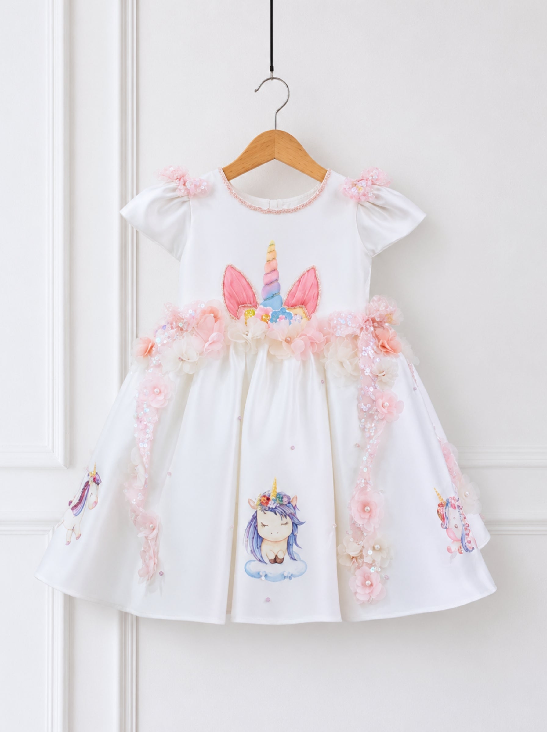 A white girls' dress with pink and multicolor unicorn prints, hanging on a wooden hanger against a white paneled wall. The dress features short puff sleeves, a floral waistband, and tulle details.