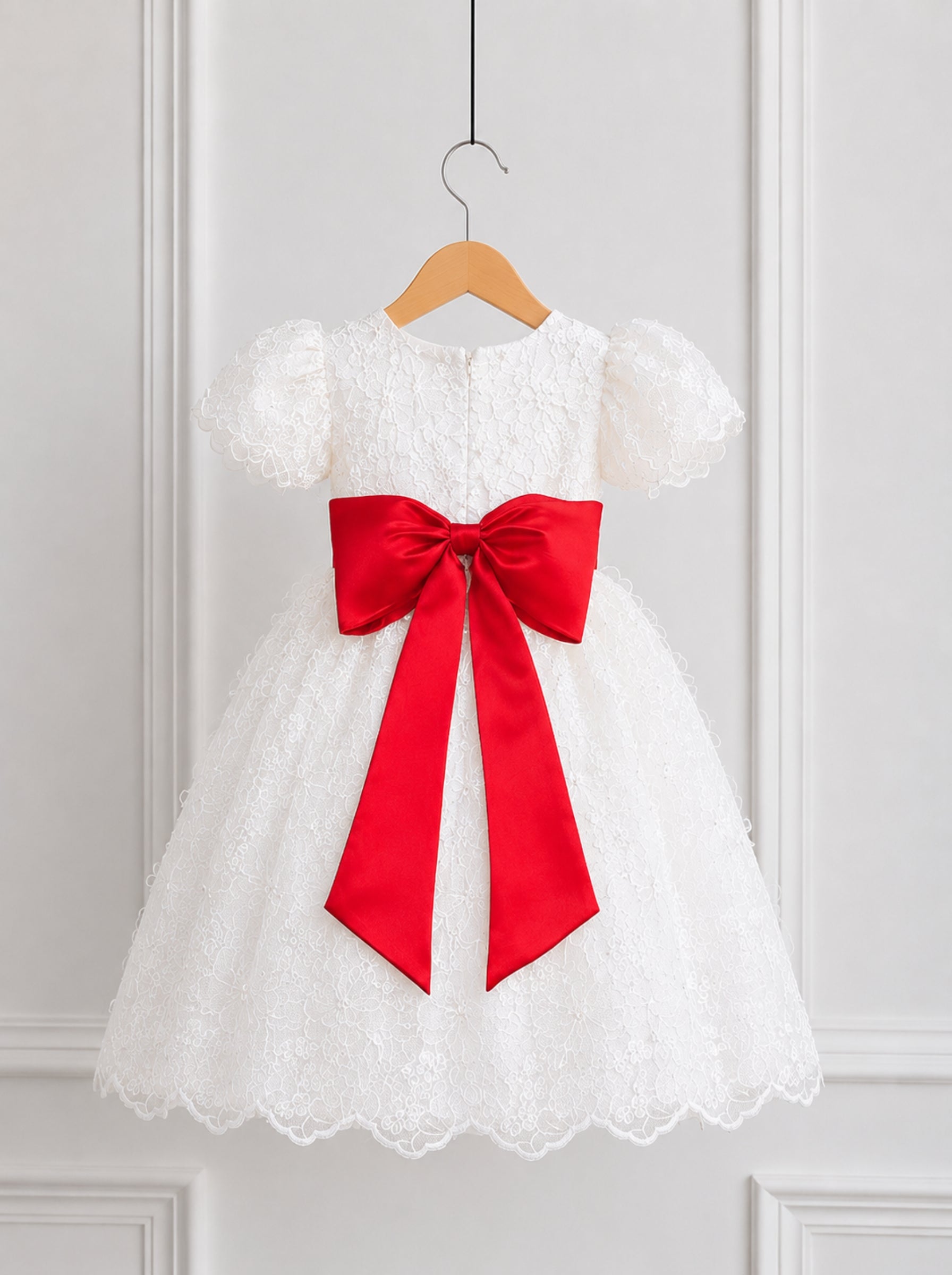 A white lace and tulle baby girl's dress with short puff sleeves and a large red satin bow at the waist, hanging on a wooden hanger against a white paneled wall.