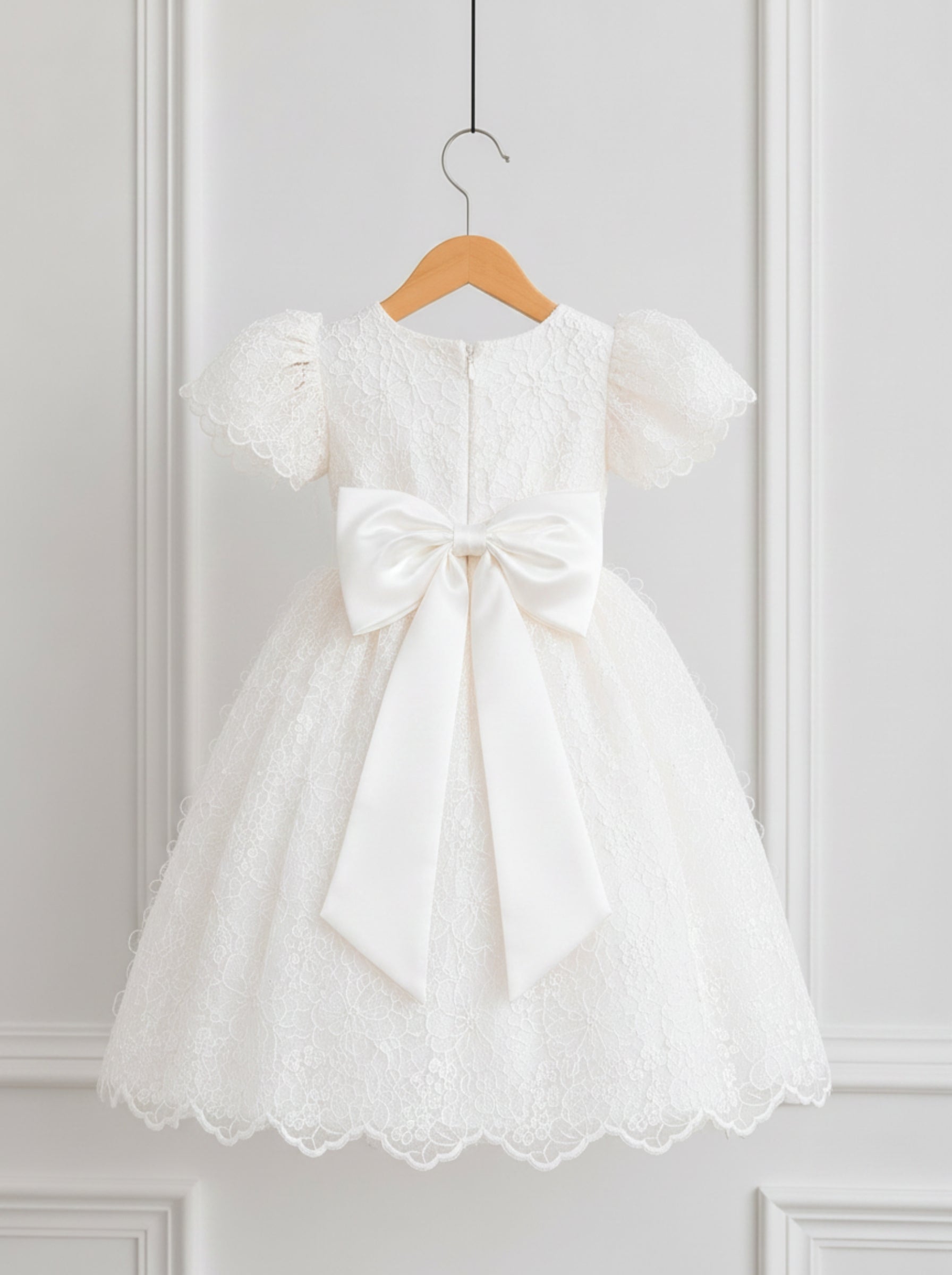 A white toddler girl's party dress with lace bodice, puff sleeves, and a large satin bow at the waist, hanging on a wooden hanger against a white paneled wall.