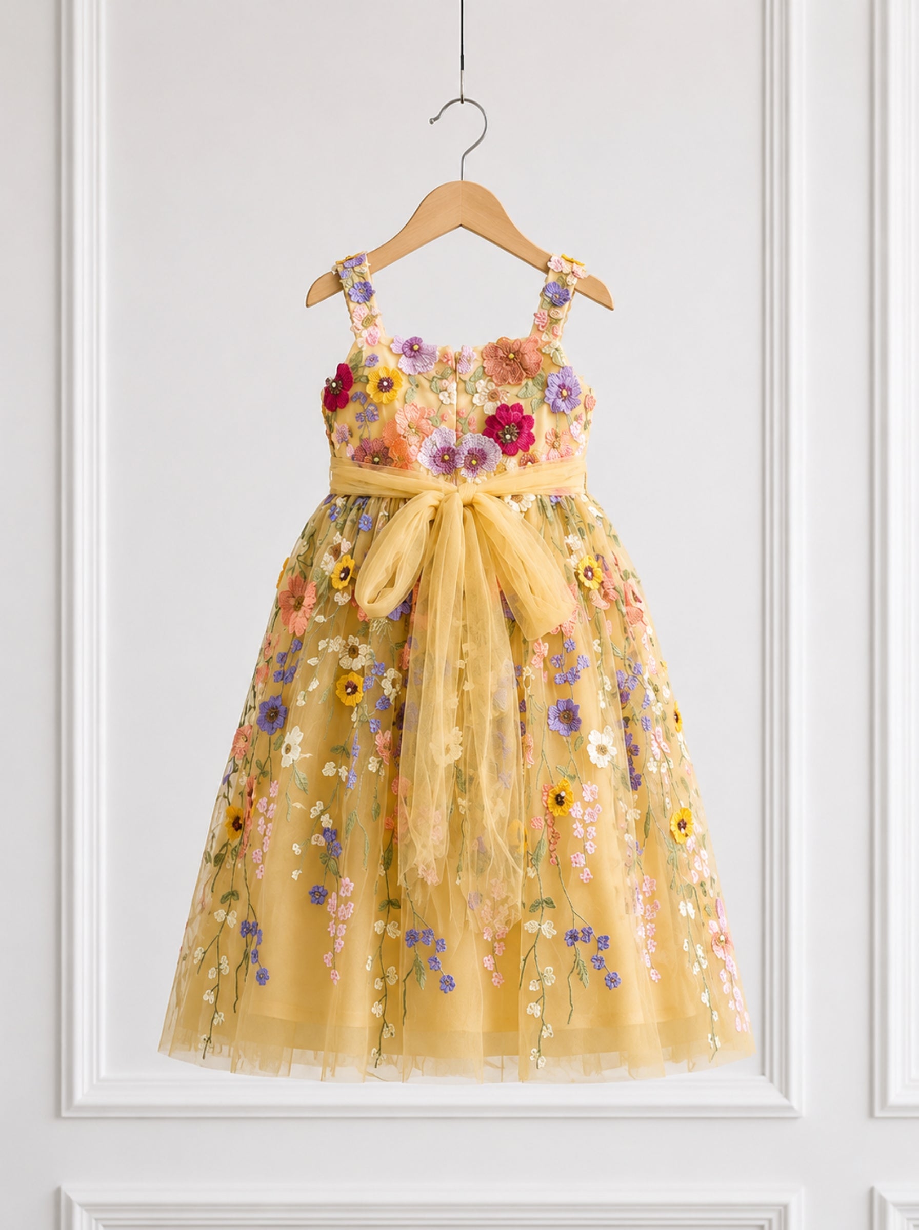 A yellow tulle dress with colorful floral embroidery and a matching yellow sash bow, hanging on a wooden hanger against a white paneled wall.
